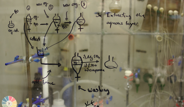 A window with scientific dry-erase writing on it and tubes and valves in the background.