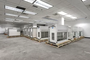 Rows of large white laboratory fume hoods sitting on wooden pallets in a brightly lit facility with a concrete floor and fluorescent ceiling lights.